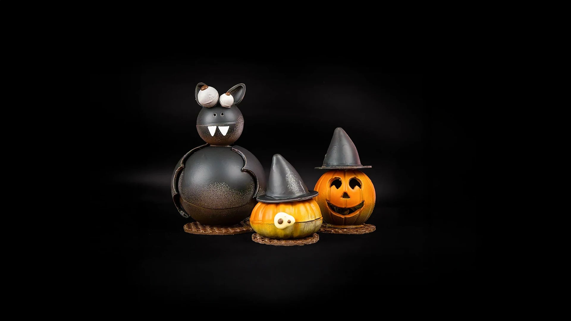 Two Halloween-themed chocolate pumpkins with hats on a black background and a chocolate Bat