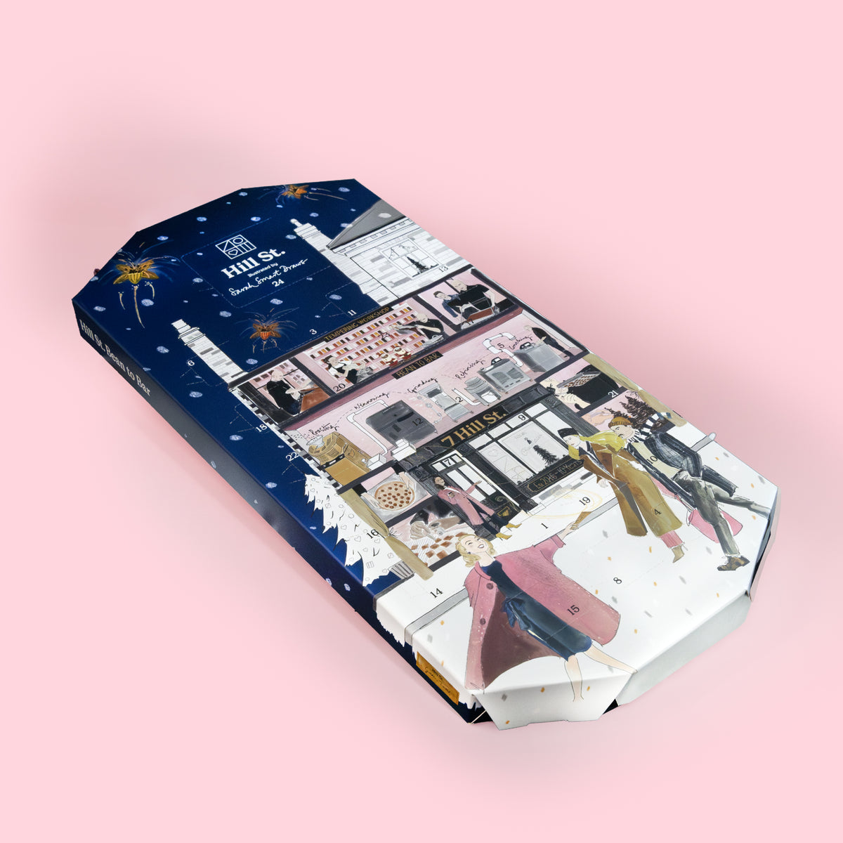 A rectangular gift box with a winter-themed collage print, featuring a dark blue snowy night sky on one side and fashion storefront scenes with people in coats on the other. The box is photographed at an angle on a solid light pink background.