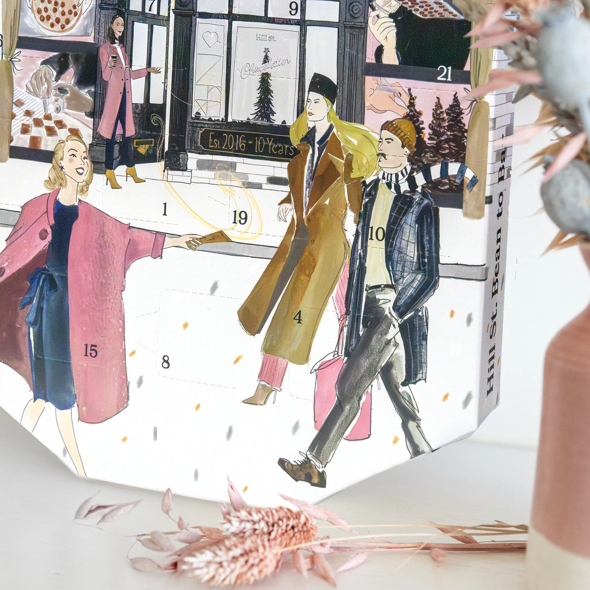 A hand-drawn fashion-themed advent calendar box on a white surface, surrounded by dried pink flowers. The box features illustrated women and men in winter coats and dresses, numbered from 1 to 21, with a Paris street scene background.