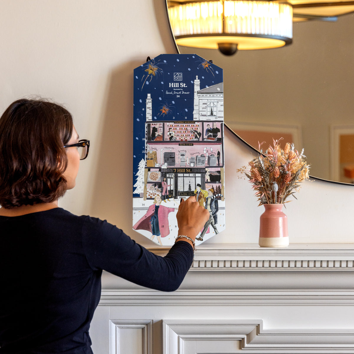 A woman in a navy long-sleeve top is shown from behind, opening a decorative illustrated advent calendar hanging on a cream-colored wall above a white mantelpiece. The calendar features a dark blue winter night design with a detailed illustration of the Hill Street Museum of Art and surrounding festive elements.