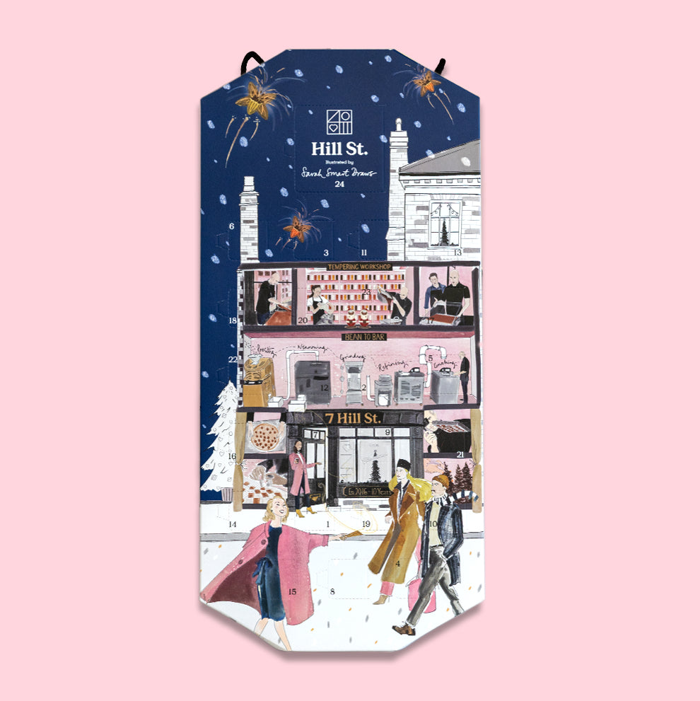 A vertical illustrated gift box or advent-style package from Hill St. with a winter city scene printed on it, shown against a solid pink background.