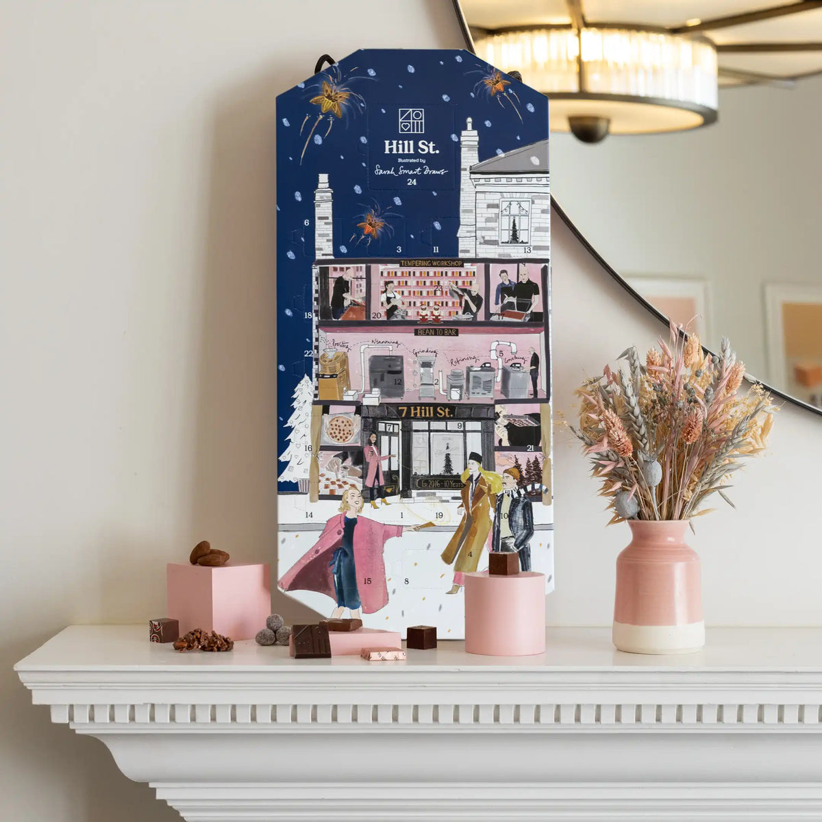 A tall illustrated gift box or advent-style package standing on a white mantelpiece, surrounded by pink boxes, chocolate pieces, a vase of dried grasses, and a mirror in a warm indoor setting. The package prominently shows the Hill St.